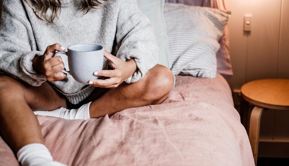 Mulher toma chá de ervas na cama, vestindo roupas confortáveis. Ambiente aconchegante e relaxante.