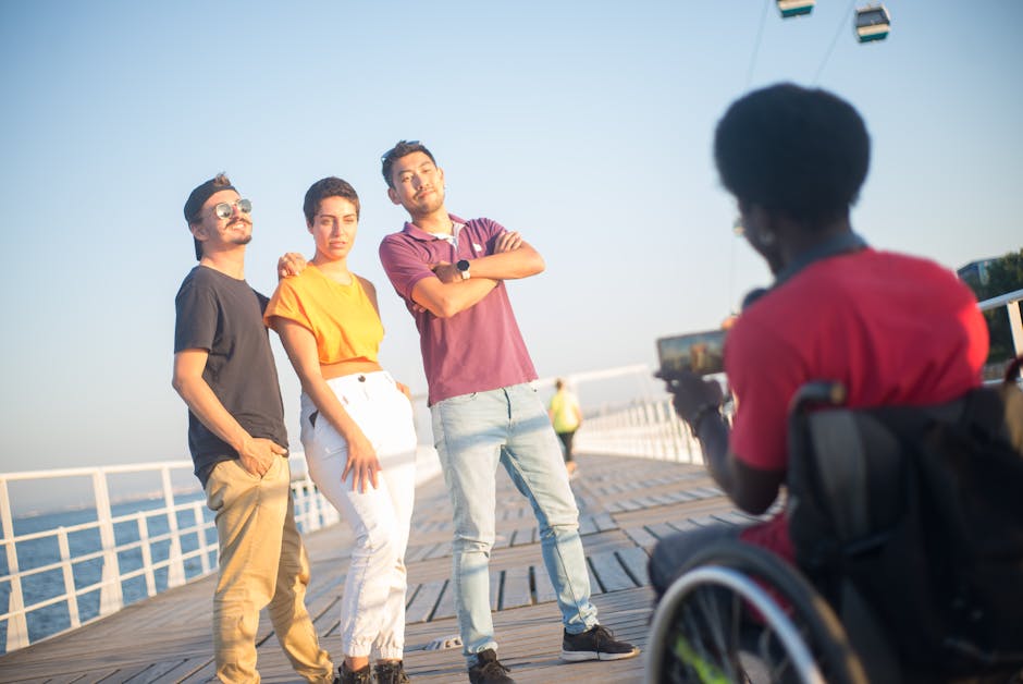 Um grupo de amigos felizes posando em uma passarela à beira-mar em um dia ensolarado, com um fotógrafo em cadeira de rodas.