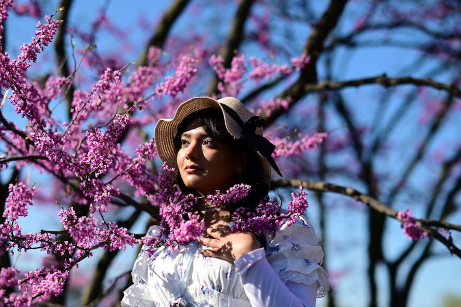 Atriz Suzy Rêgo em meio a flores cor-de-rosa, vestindo traje vintage, sob céu azul claro.