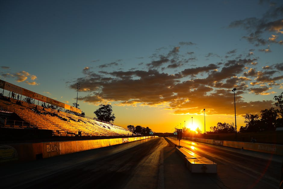 Pista de corrida vazia ao pôr do sol, com arquibancadas e céu colorido refletidos no asfalto.